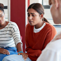 Image of 3 women in a group therapy type environment. The woman in th center is crying while the other two offer comfort.