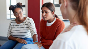 Image of 3 women sitting together in a group therapy environment. The middle woman is crying.