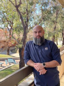 Bearded man leaning on a wooden fence outdoors.