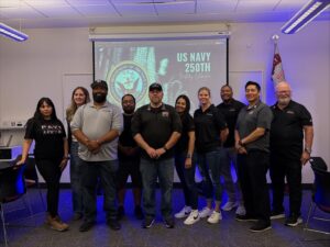 10 members of the UNLV PAVE team posed in front of a projector screen reading" US Navy 250th"