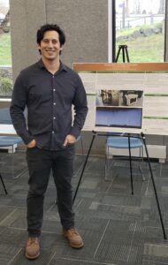 Figure standing in front of a poster presentation, smiling with hands in pockets.