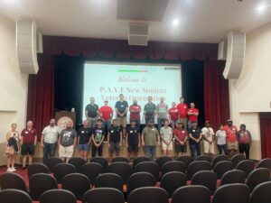 Group of people standing in 2 lines on a stage in front of a projector screen reading: "PAVE New Student Veteran Orientation"